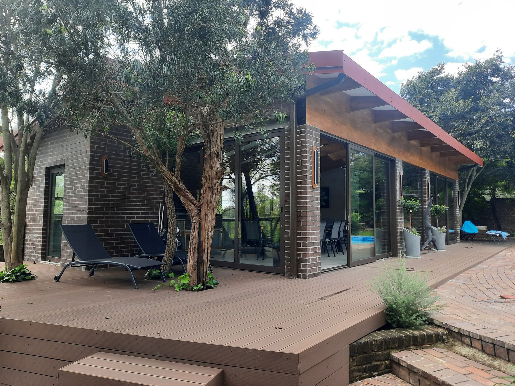 Timber-decked entertainment pavilion in Bryanston with a red-edged mono-pitch roof, built around an existing olive tree
